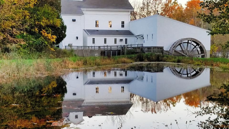 A white building with a large water wheel and a reflection in a calm pond.