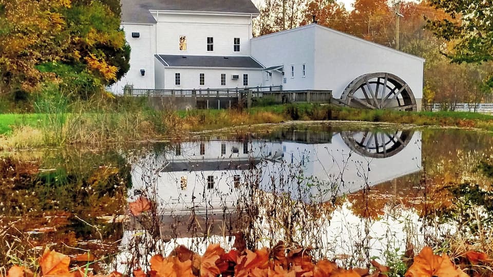 A white building with a large water wheel in a pond surrounded by autumn leaves.
