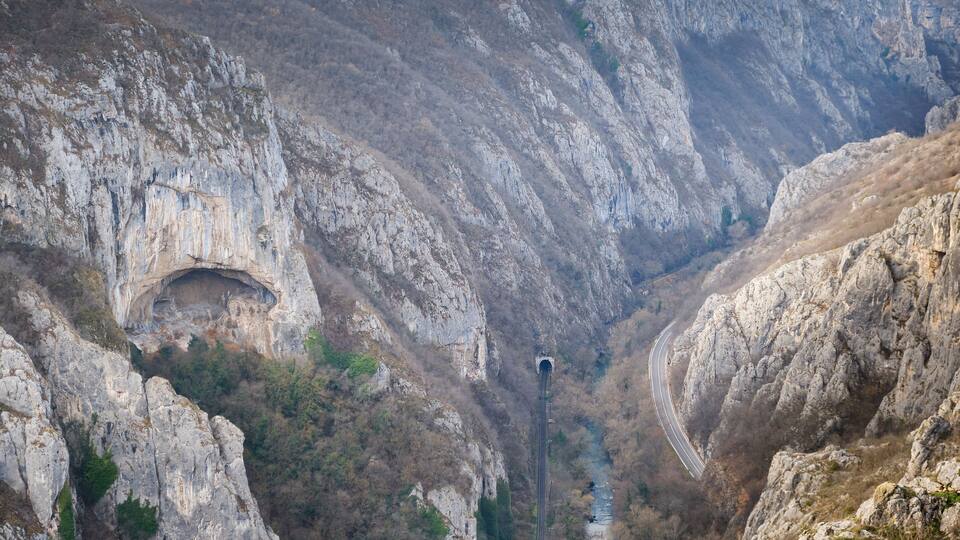 A mountainous landscape with a narrow road winding through a rocky gorge, a tunnel entrance, and a river flowing through the valley.