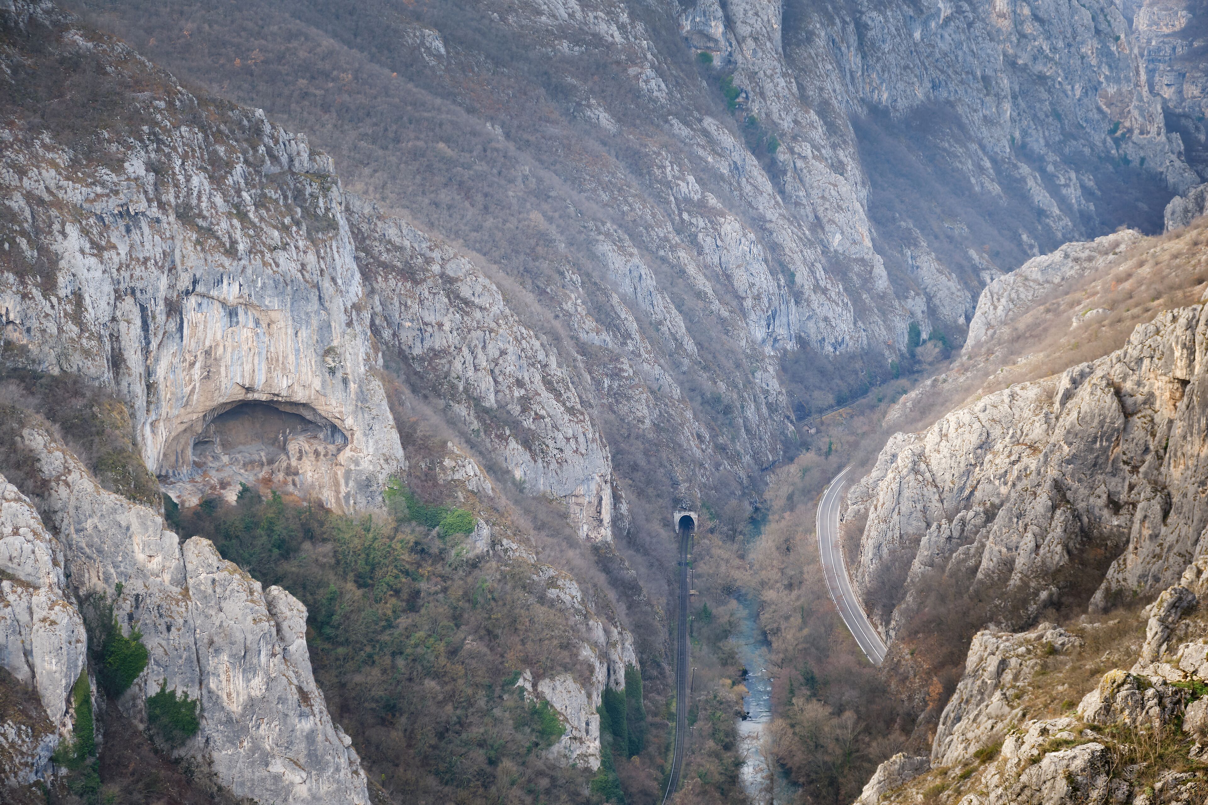 A mountainous landscape with a narrow road winding through a rocky gorge, a tunnel entrance, and a river flowing through the valley.