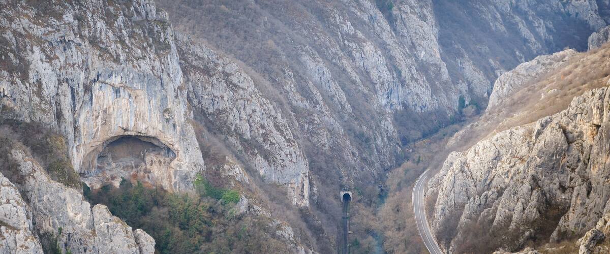 A mountainous landscape with a narrow road winding through a rocky gorge, a tunnel entrance, and a river flowing through the valley.