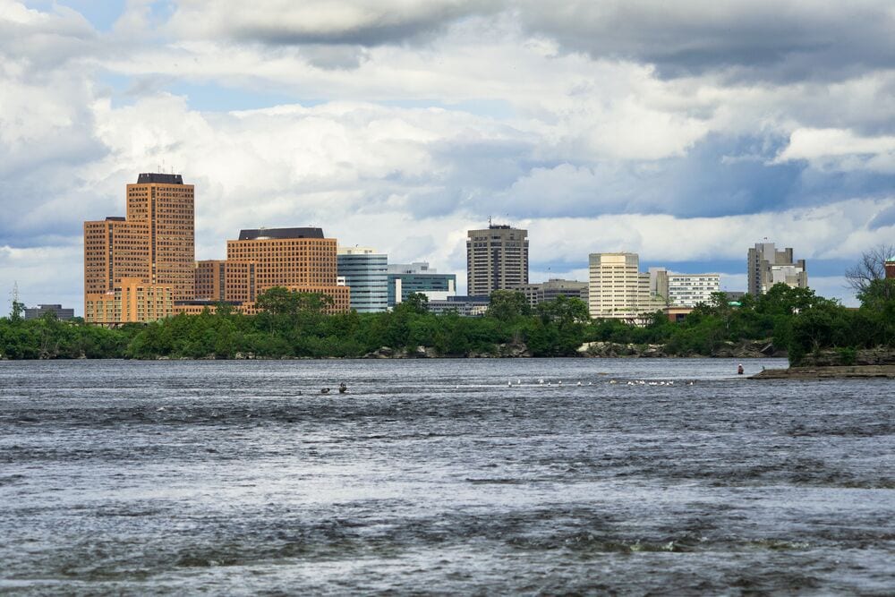 A city skyline with several high-rise buildings, a river in the foreground, and a cloudy sky.