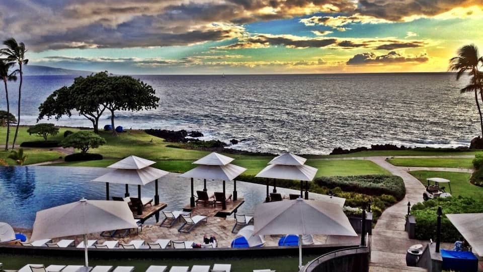Zwembadgedeelte van een resort met ligbedden en parasols, met uitzicht op de oceaan en een bewolkte lucht.