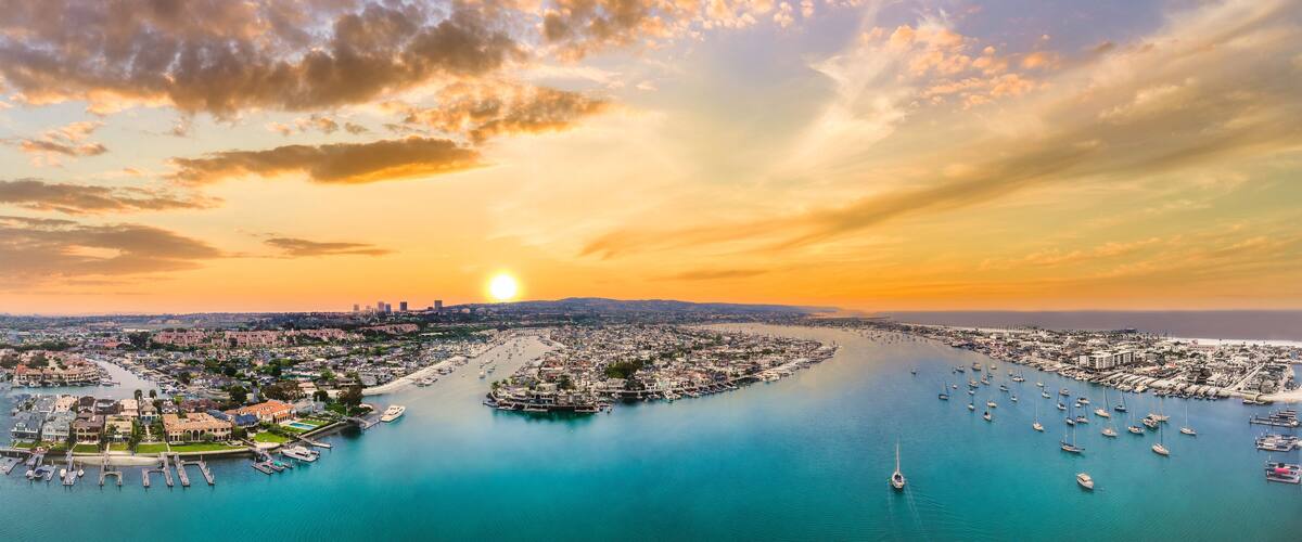 A coastal cityscape with a marina filled with boats, a river running through the city, and a sunset over the horizon.