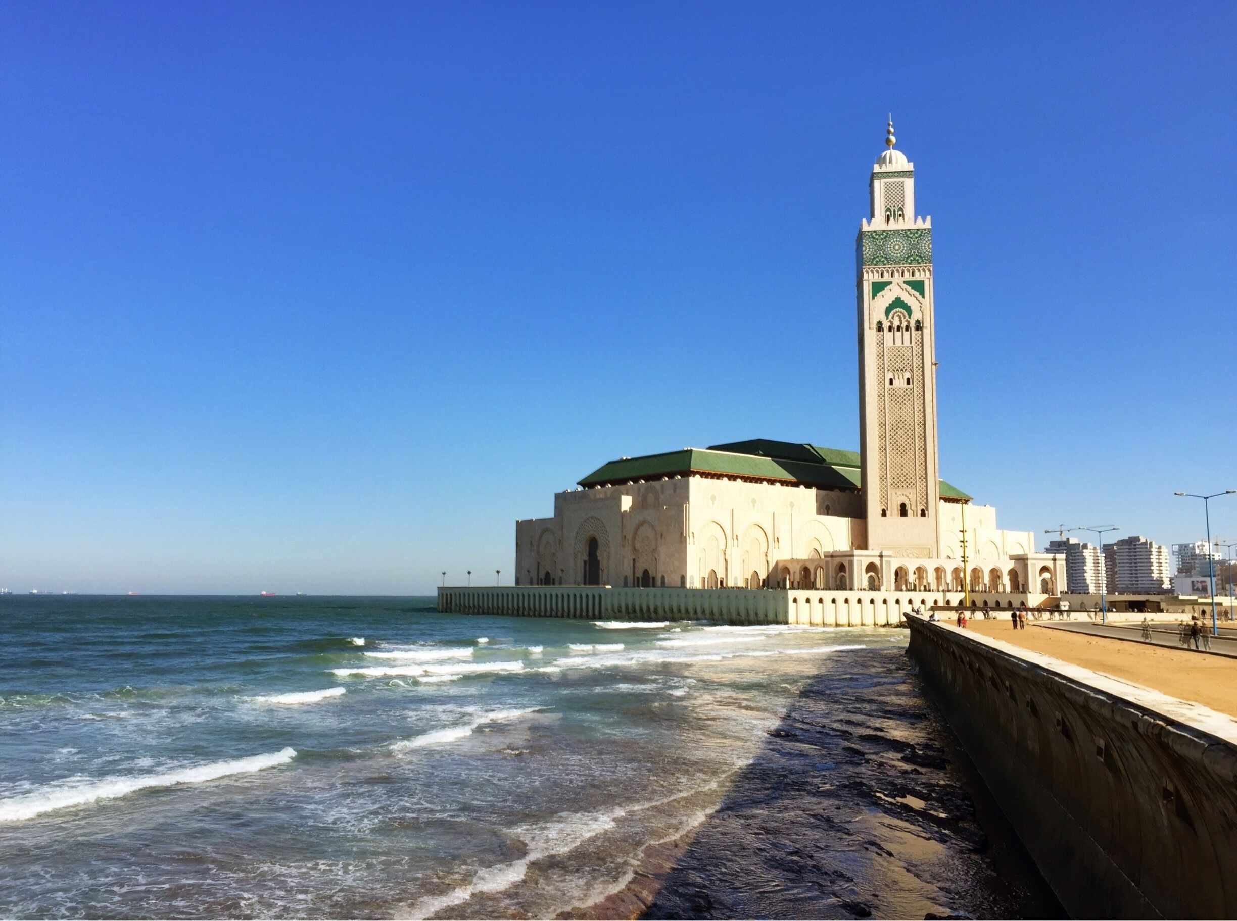 A coastal mosque with a tall minaret and a green dome, adjacent to a sandy beach and the sea.