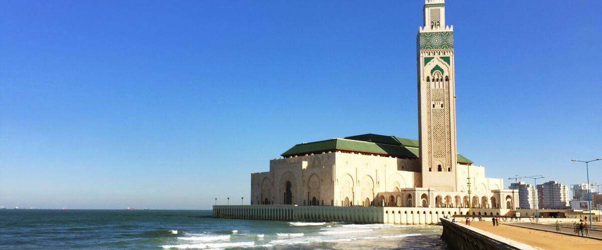 A coastal mosque with a tall minaret and a green dome, adjacent to a sandy beach and the sea.
