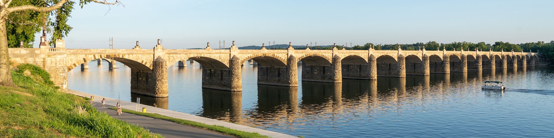 A stone bridge over a calm river, with a paved walkway on the left and trees on the right.