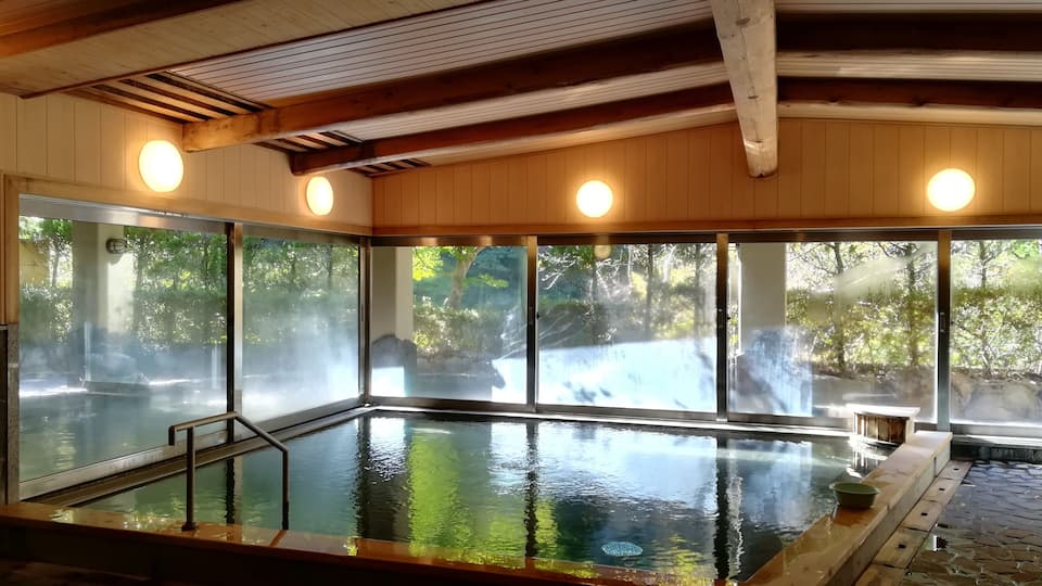 A large indoor swimming pool with glass walls, wooden ceiling beams, and a stone-tiled floor.