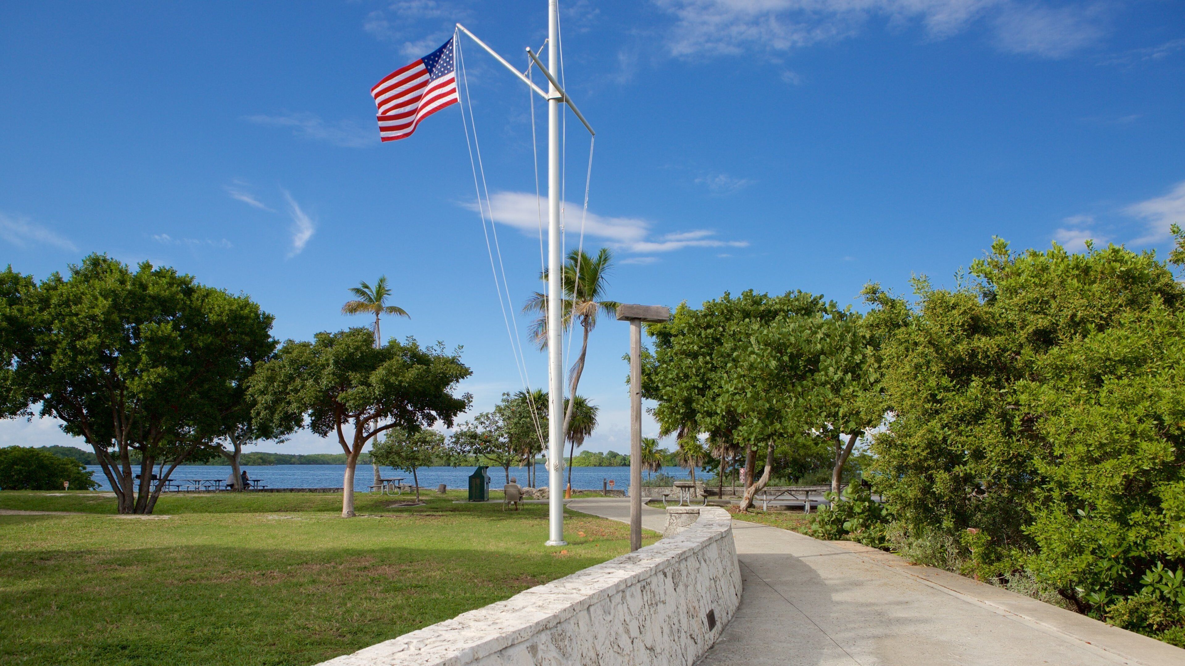 A park with a flagpole, trees, a pathway, and a body of water in the background.