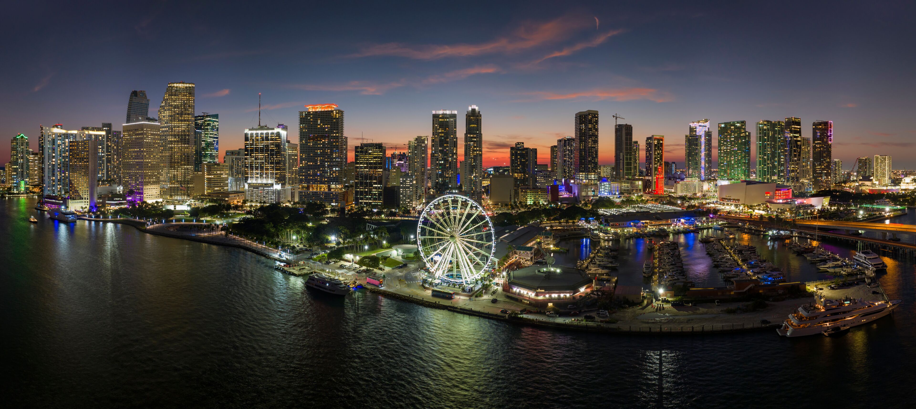 A city skyline at dusk with illuminated skyscrapers, a Ferris wheel, and a marina with boats.