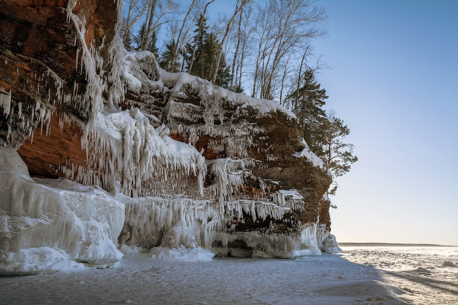 A frozen cliff with icicles and snow-covered trees.