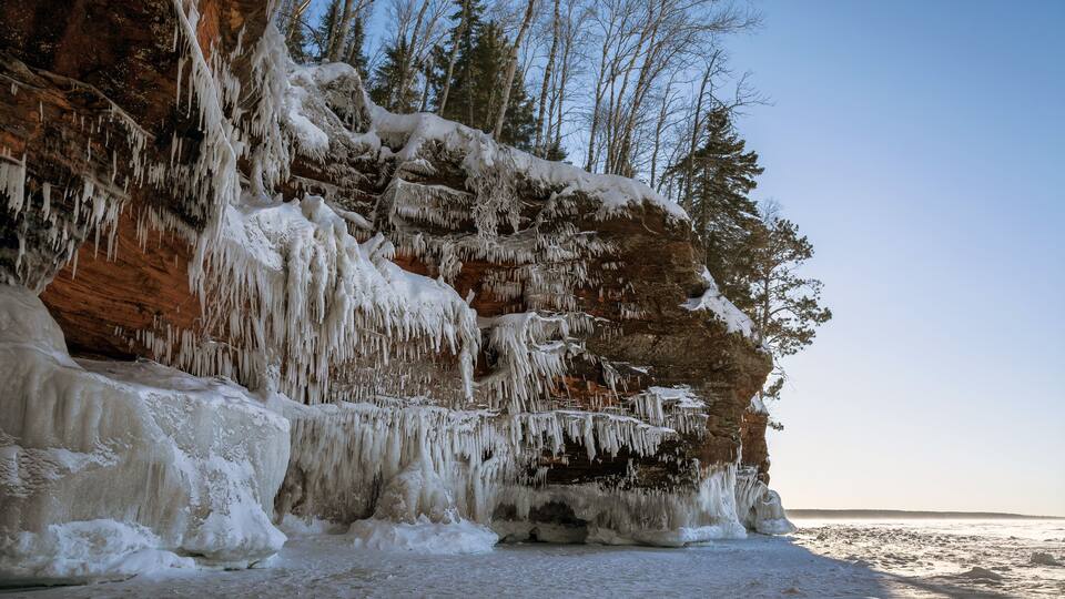 A frozen cliff with icicles and snow-covered trees.