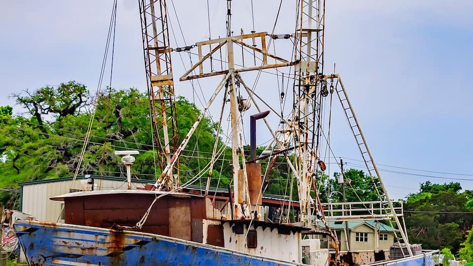 A rusted, blue fishing boat docked at a pier with a green landscape in the background.