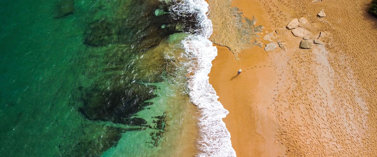 An aerial view of a beach with clear turquoise water, a sandy shore, and scattered rocks.
