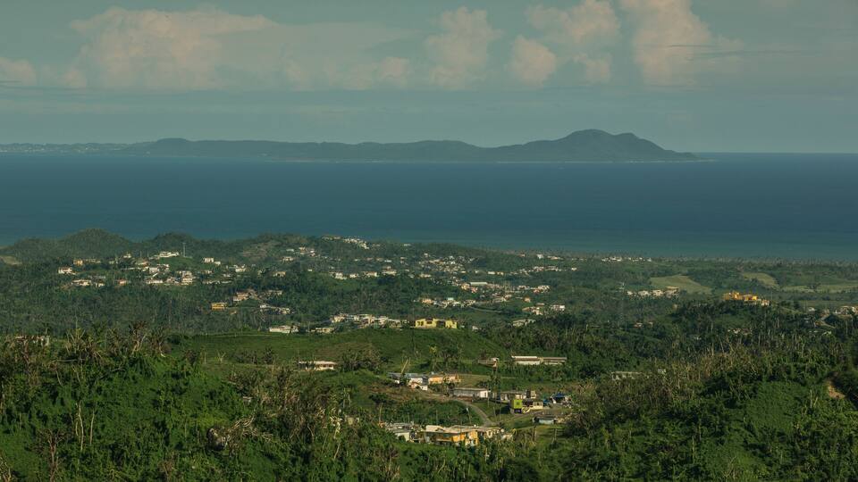 A coastal town with houses and greenery, a mountain in the distance, and the ocean.