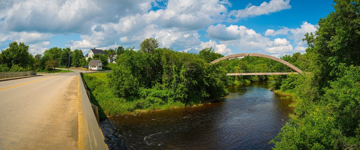 A river with a wooden bridge, surrounded by lush greenery and houses.