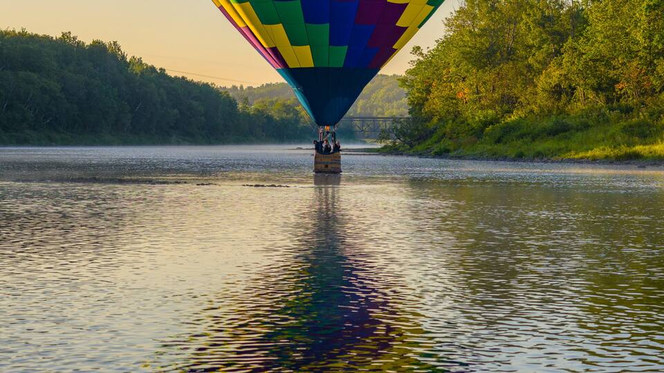 A hot air balloon over a calm river with a forested riverbank.