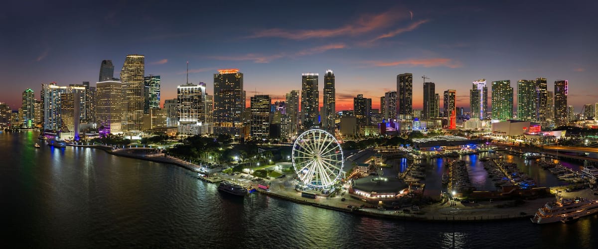 A city skyline at night with illuminated buildings, a Ferris wheel, and a waterfront marina.