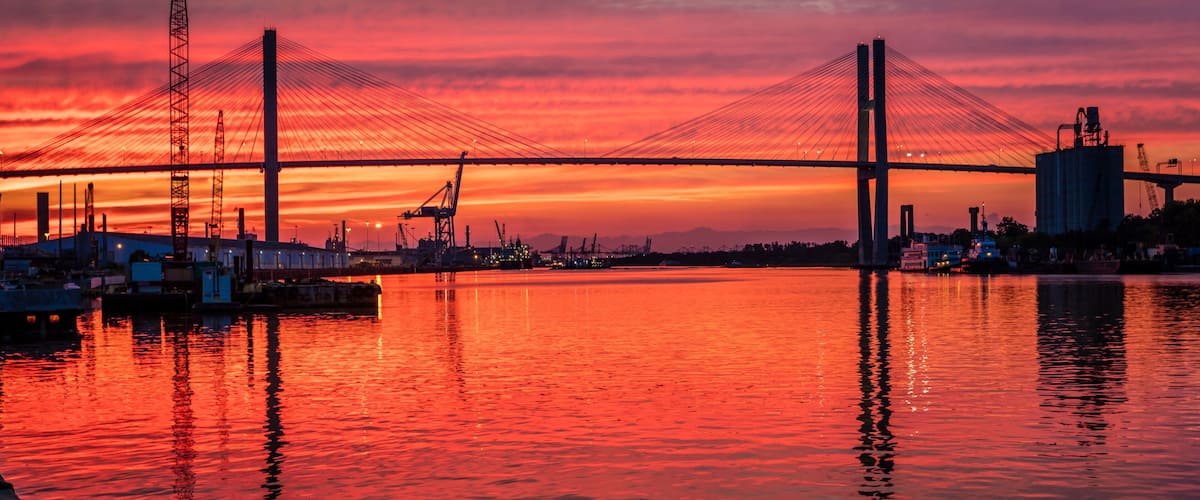 A bridge at sunset with a vibrant sky, calm water, and industrial structures in the background.