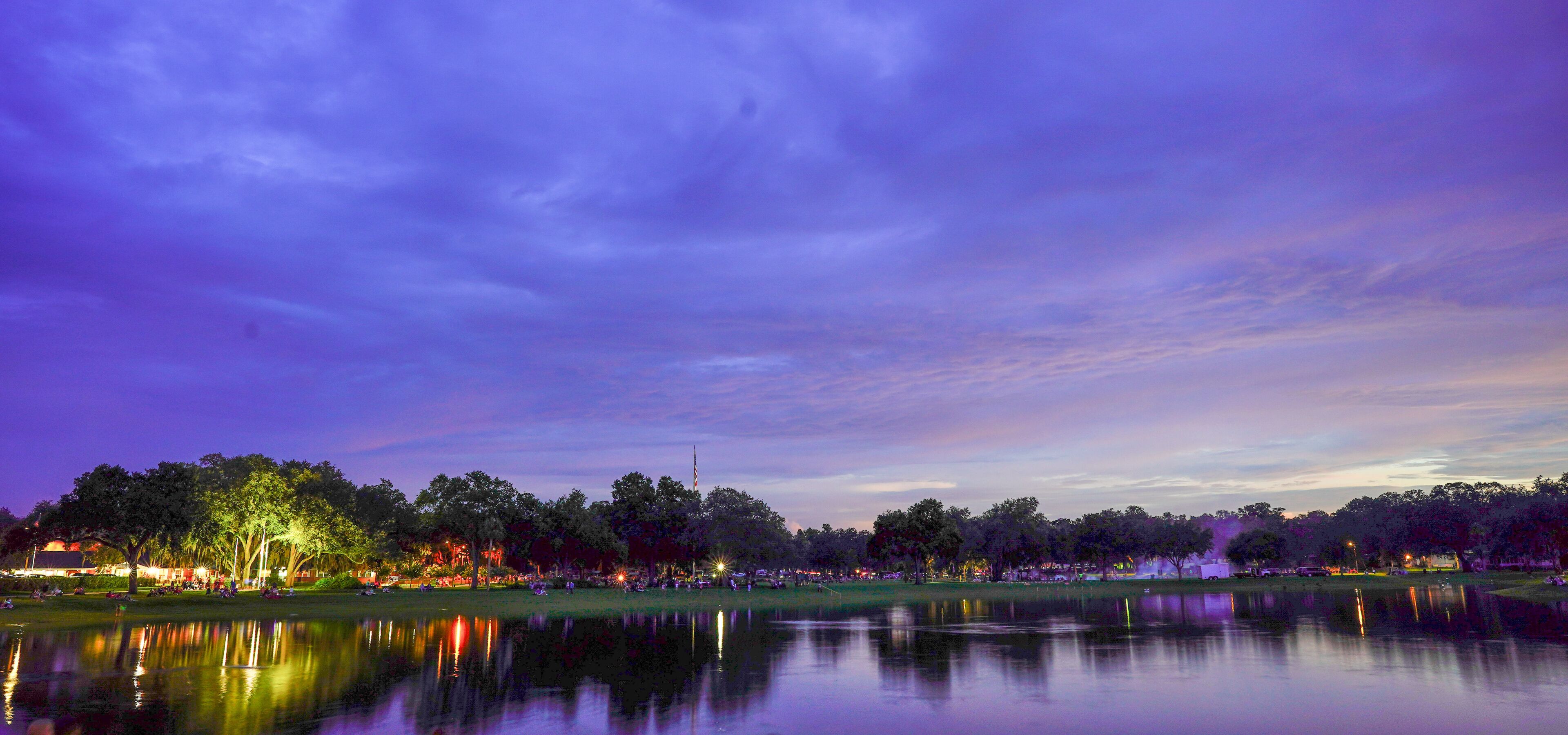 A calm lake at dusk with trees and distant lights reflecting on the water.