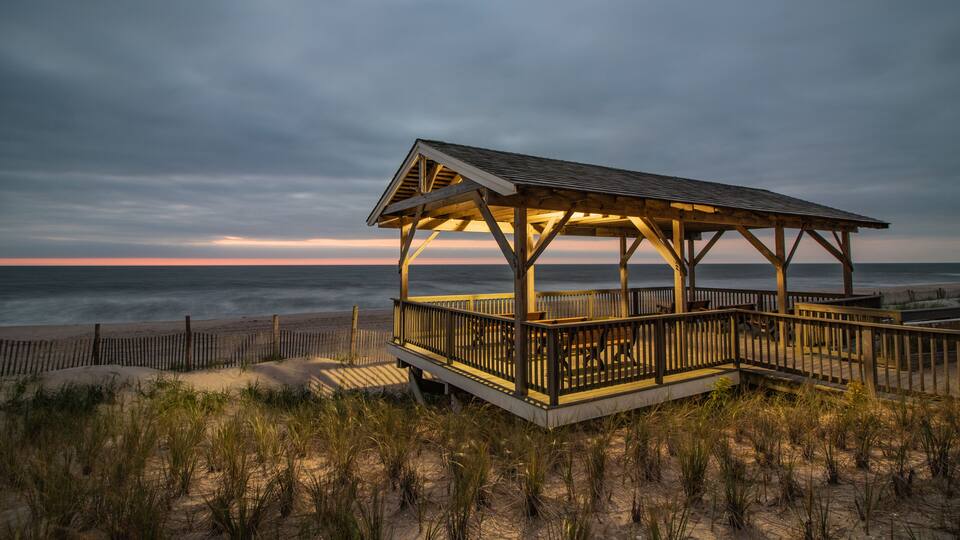 A wooden beach pavilion with a metal roof, surrounded by sand dunes and beach grasses.
