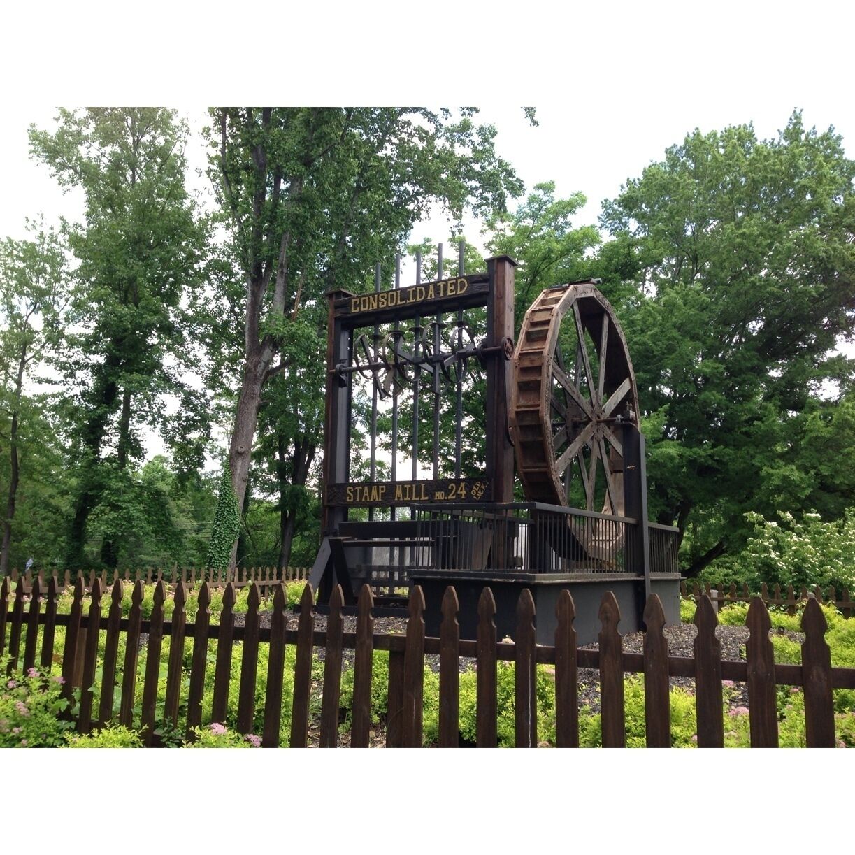 A historic water wheel labeled "CONSOLIDATED STAMP MILL No. 24" surrounded by a wooden fence and lush greenery.