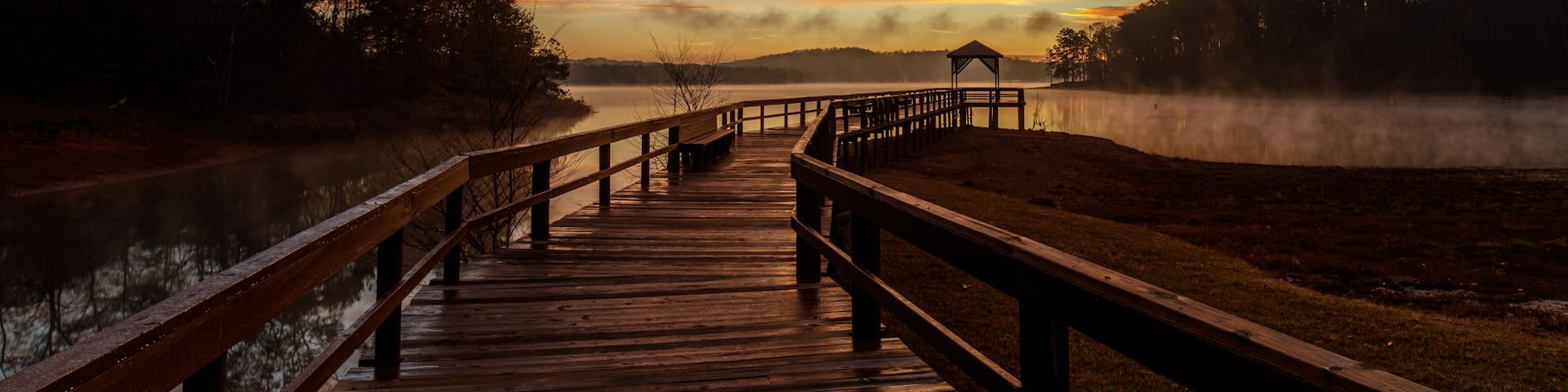 A wooden boardwalk leading to a gazebo by a lake with a sunset sky.
