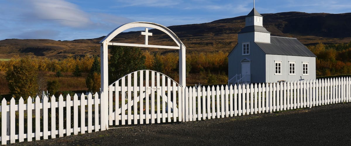 A white church with a cross on the roof, surrounded by a white picket fence, set against a backdrop of hills and autumn foliage.