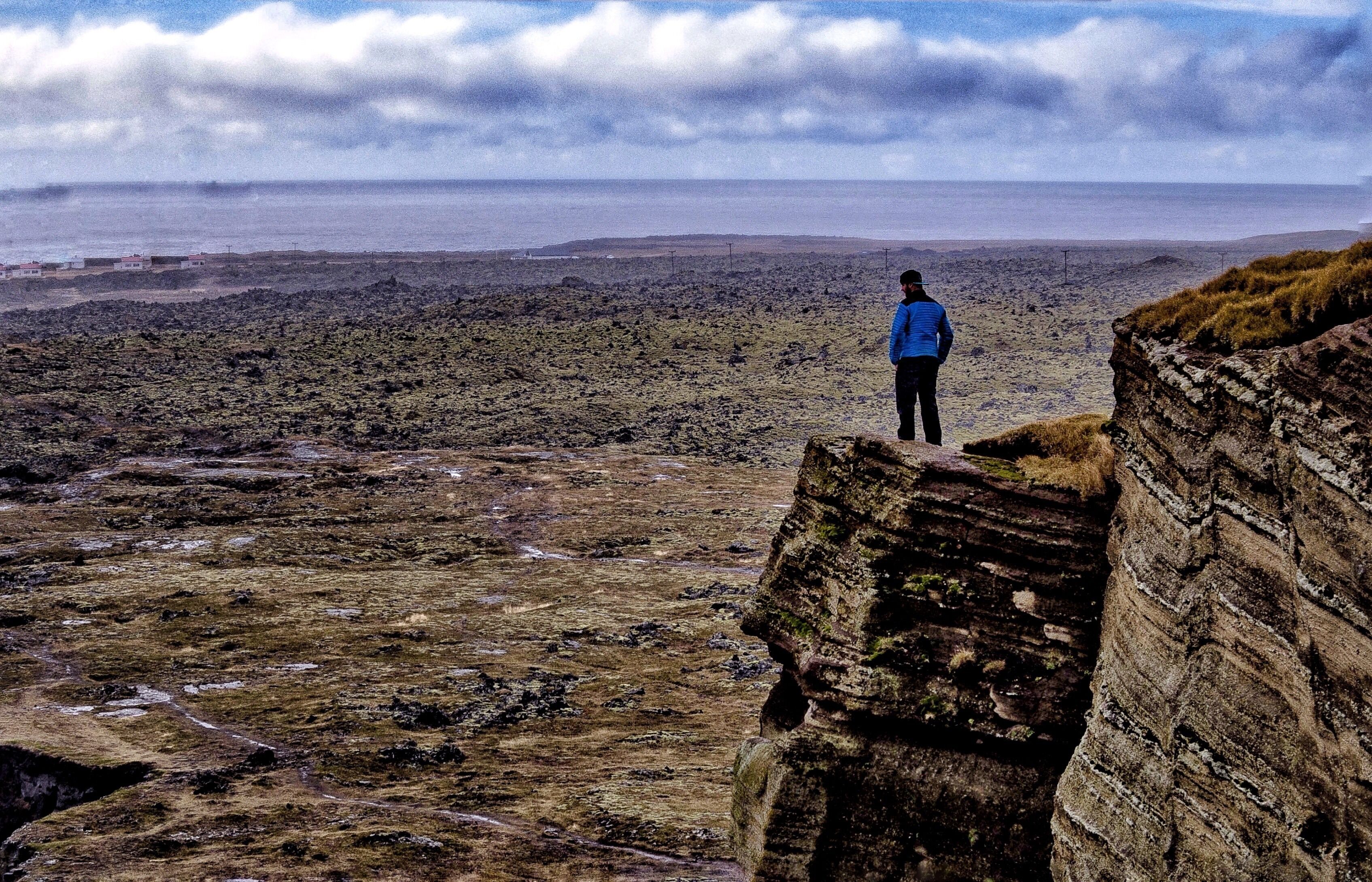 A person standing on a rocky outcrop overlooking a barren landscape with distant hills and a cloudy sky.