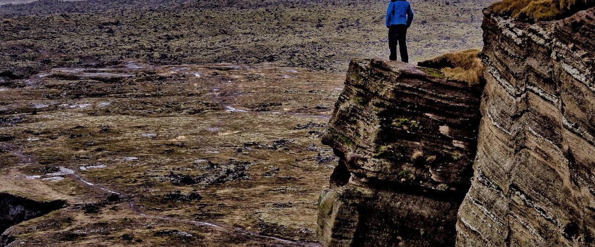 A person standing on a rocky outcrop overlooking a barren landscape with distant hills and a cloudy sky.