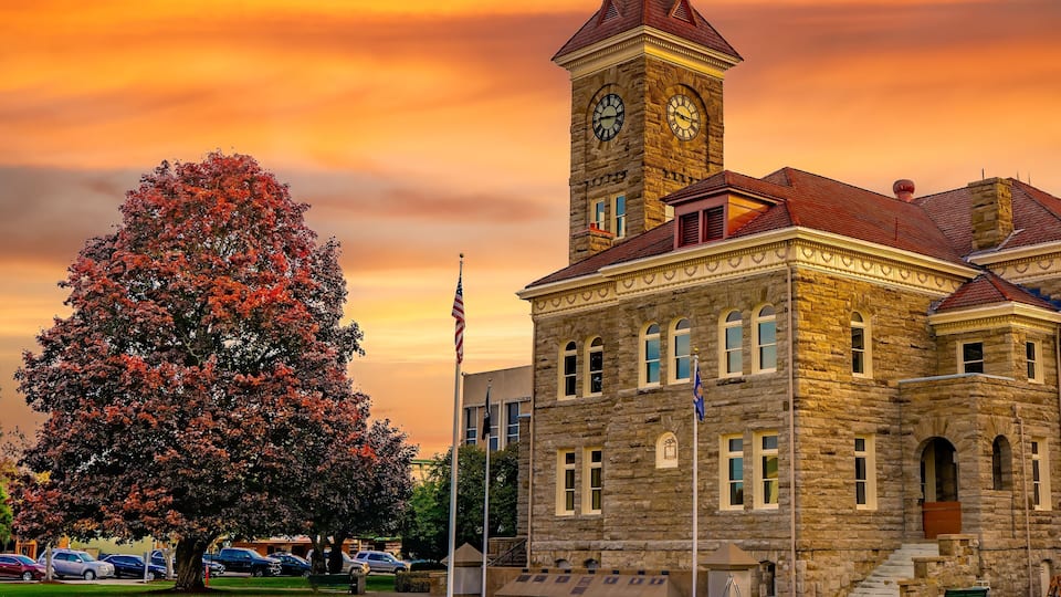 A historic stone building with a clock tower, surrounded by a park with a large tree and a flagpole.
