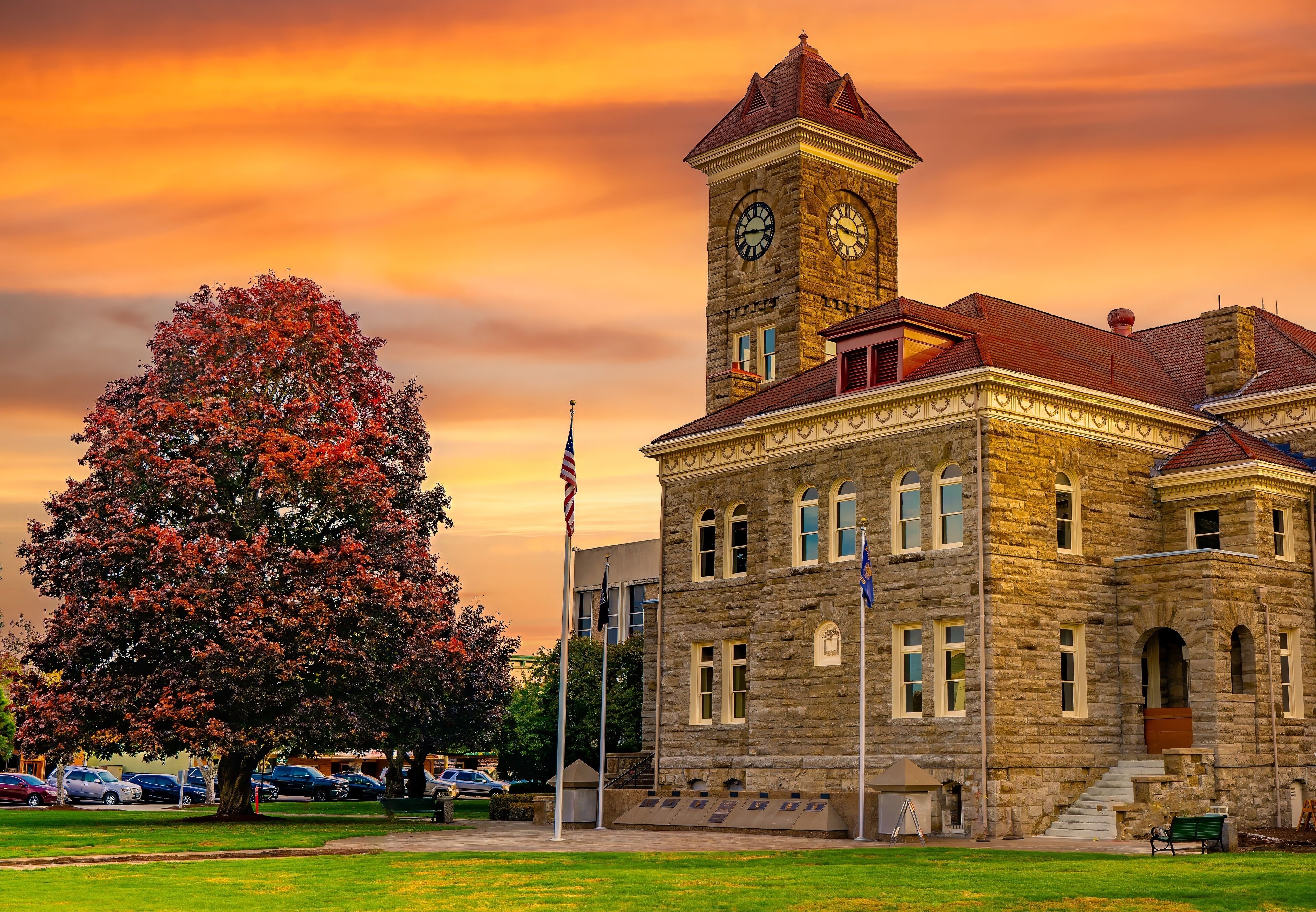 A historic stone building with a clock tower, surrounded by a park with a large tree and a flagpole.