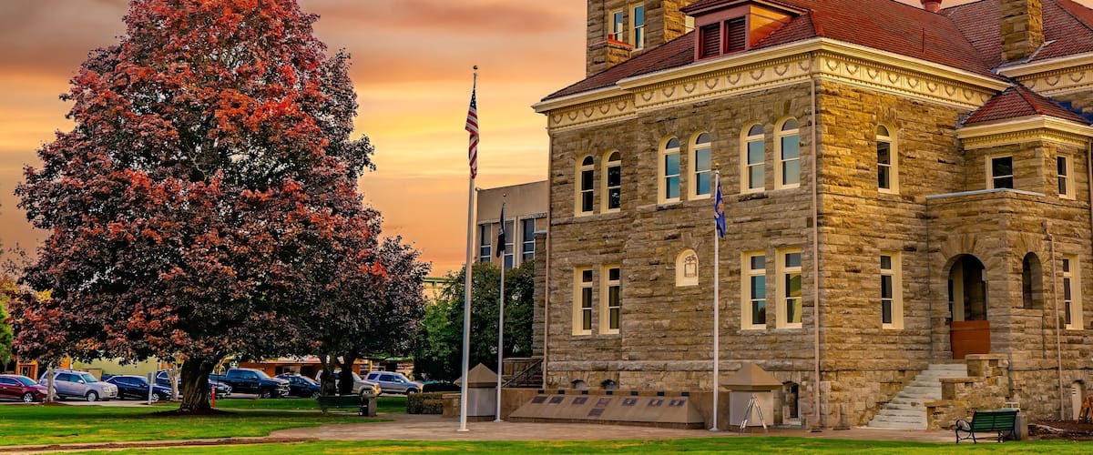 A historic stone building with a clock tower, surrounded by a park with a large tree and a flagpole.