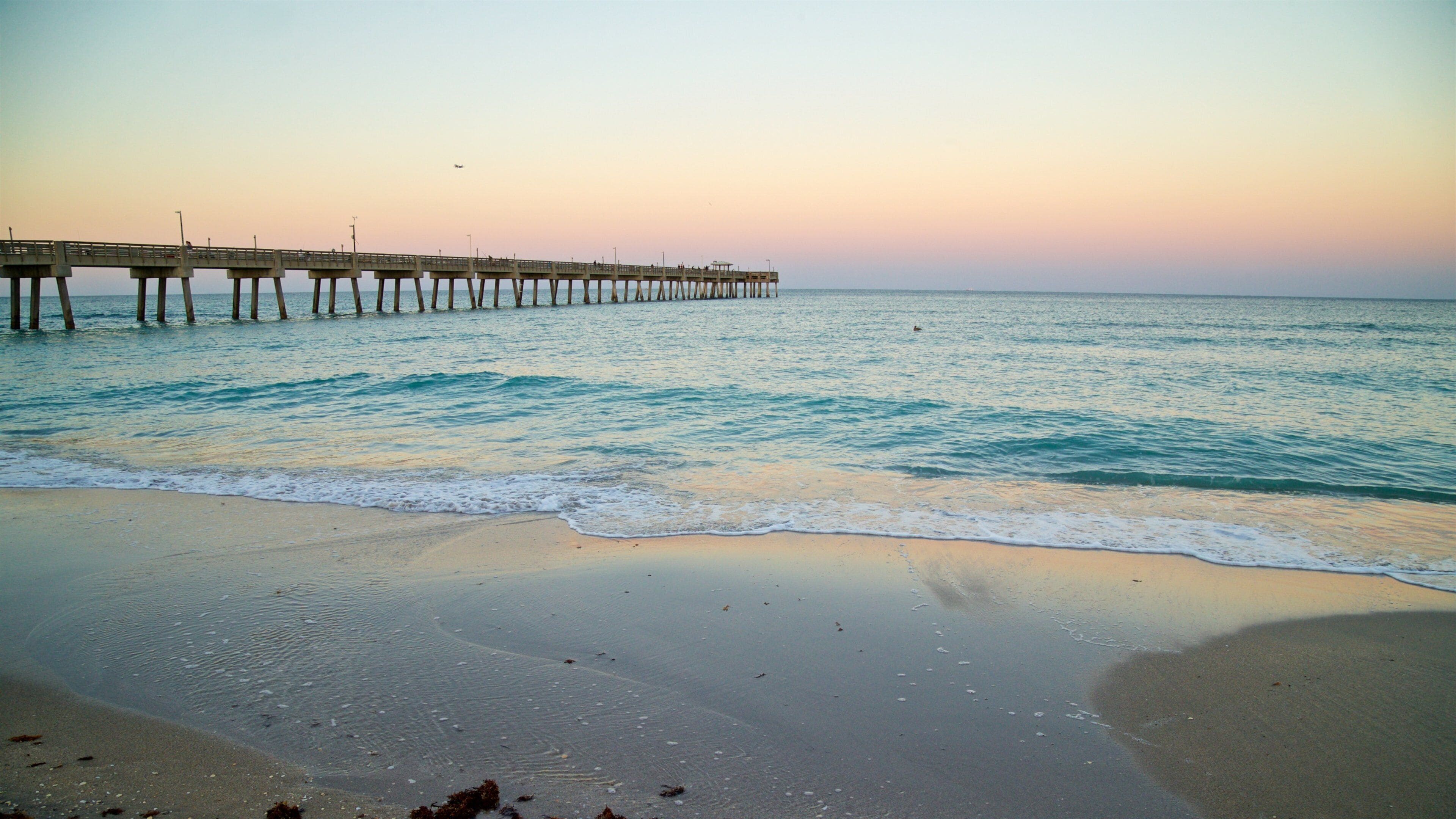 A beach with waves, a pier extending into the water, and a sunset sky.