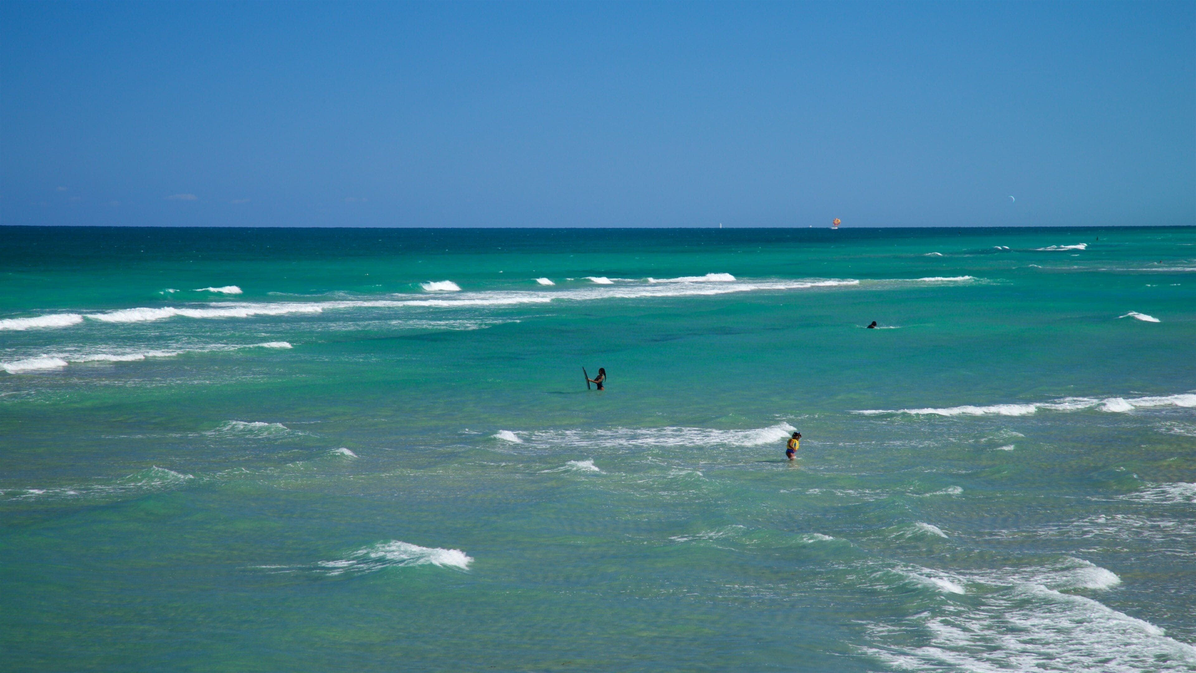 A clear turquoise sea with gentle waves, a distant lighthouse, and a few people in the water.