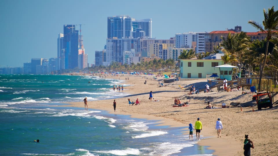 A beach with people walking along the shore, palm trees, and a city skyline in the background.