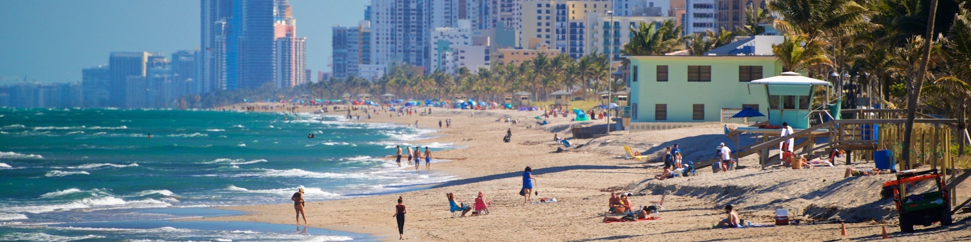 A beach with people walking along the shore, palm trees, and a city skyline in the background.