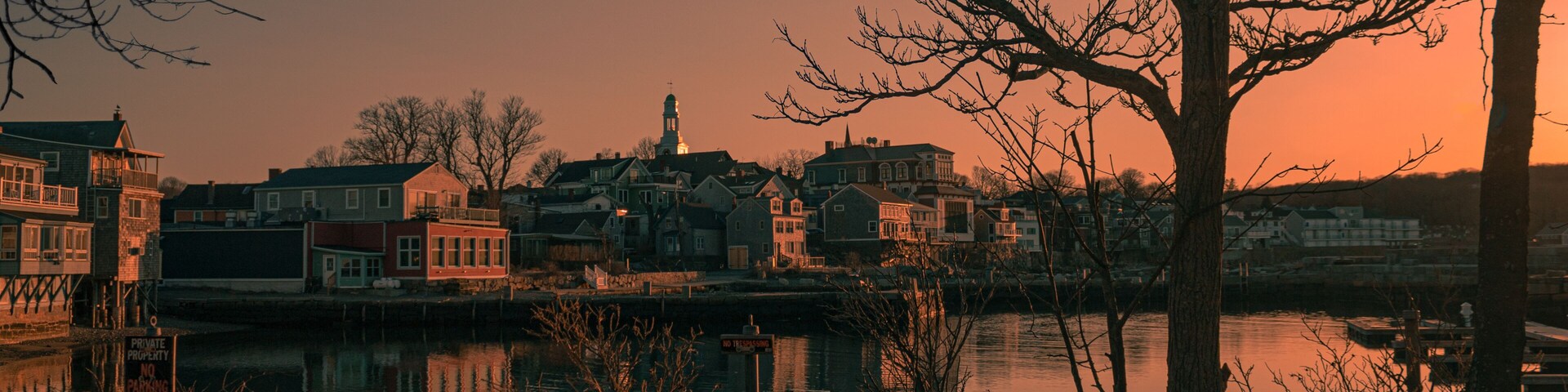 Uma cidade à beira-rio, com edifícios históricos, uma imponente torre de igreja e um rio calmo refletindo o pôr do sol.