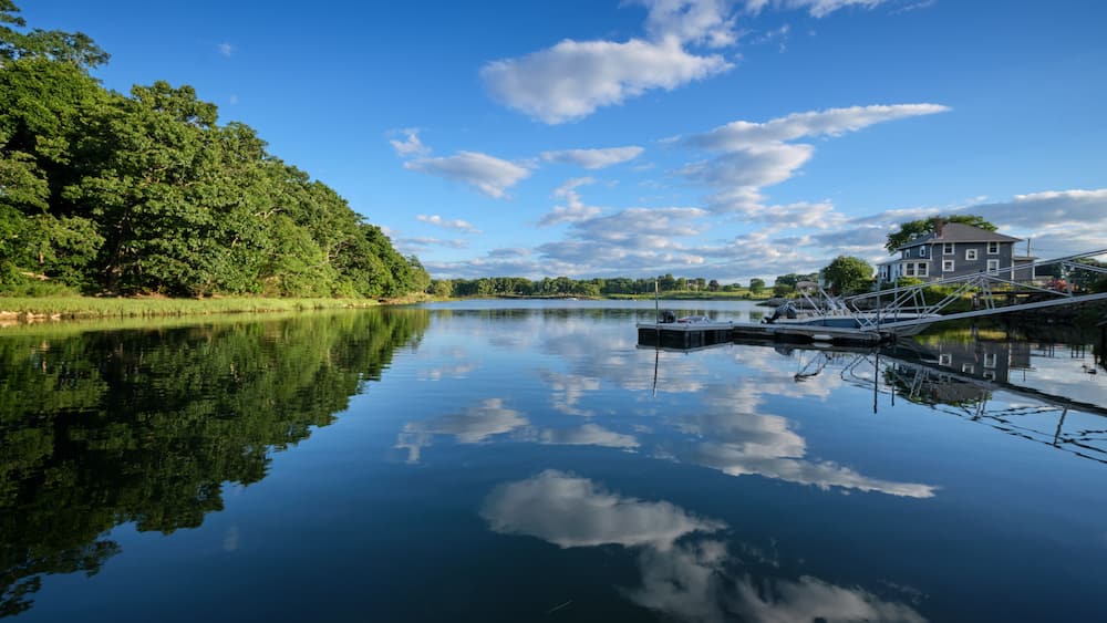Ein ruhiger See mit einem Steg, ein Haus auf der rechten Seite und dichte Bäume links.