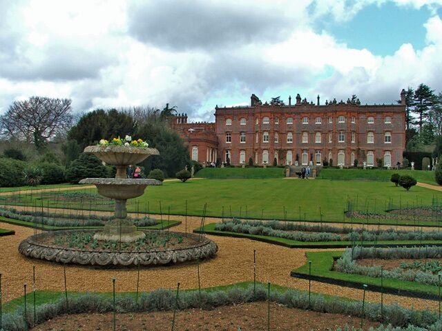 A formal garden with a central fountain, neatly trimmed hedges, and a large red building in the background.