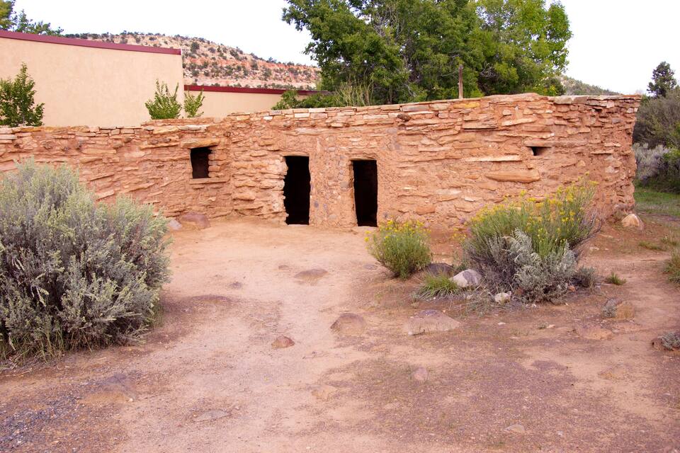 Un bâtiment en pierre, doté de deux portes et d’un toit rouge, entouré de la végétation typique du désert.