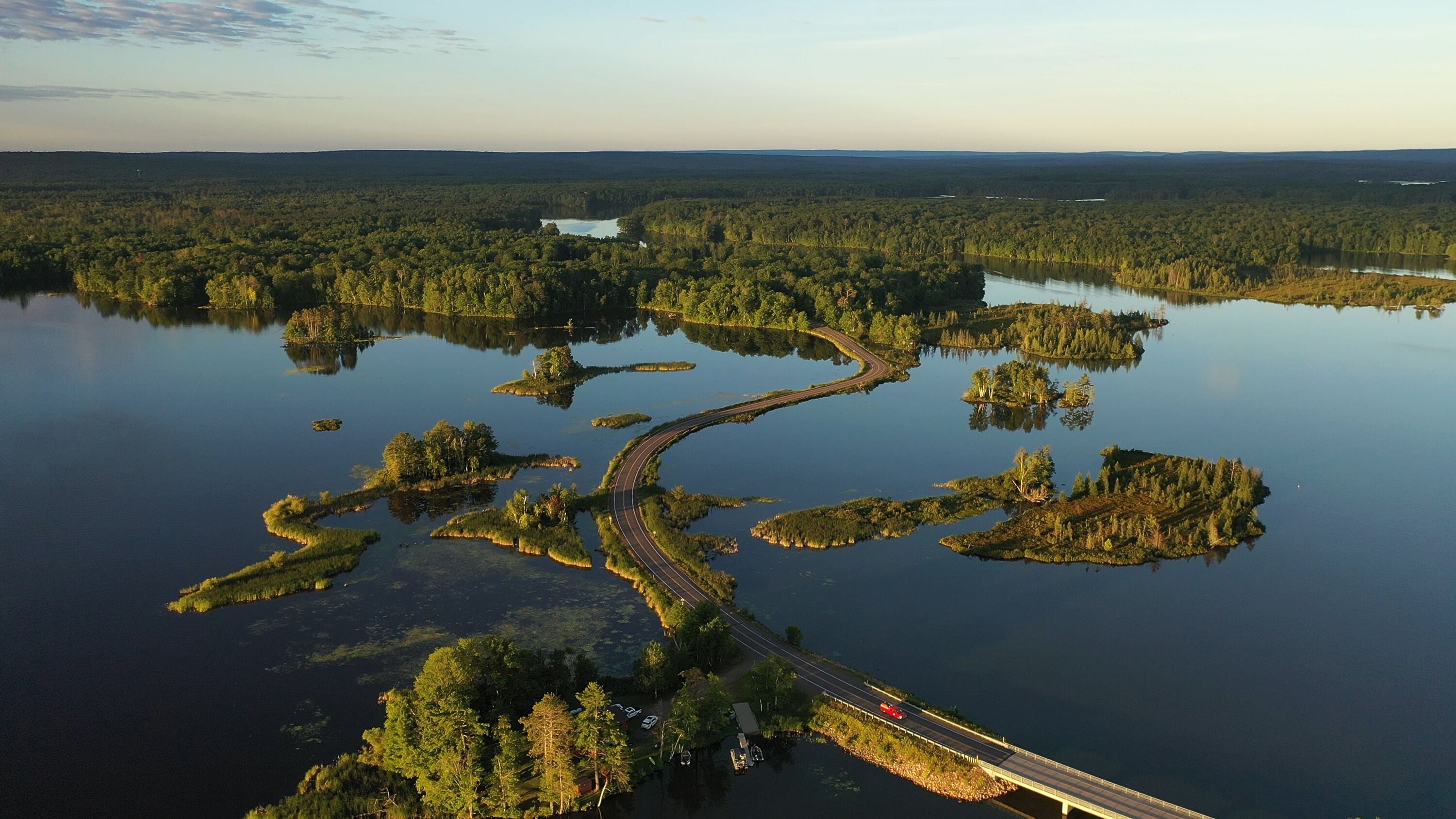 Establishing shot of american wilderness. Aerial view of scenic road through lake, and wooded islands. Calm morning water, dawn sky, view from above.