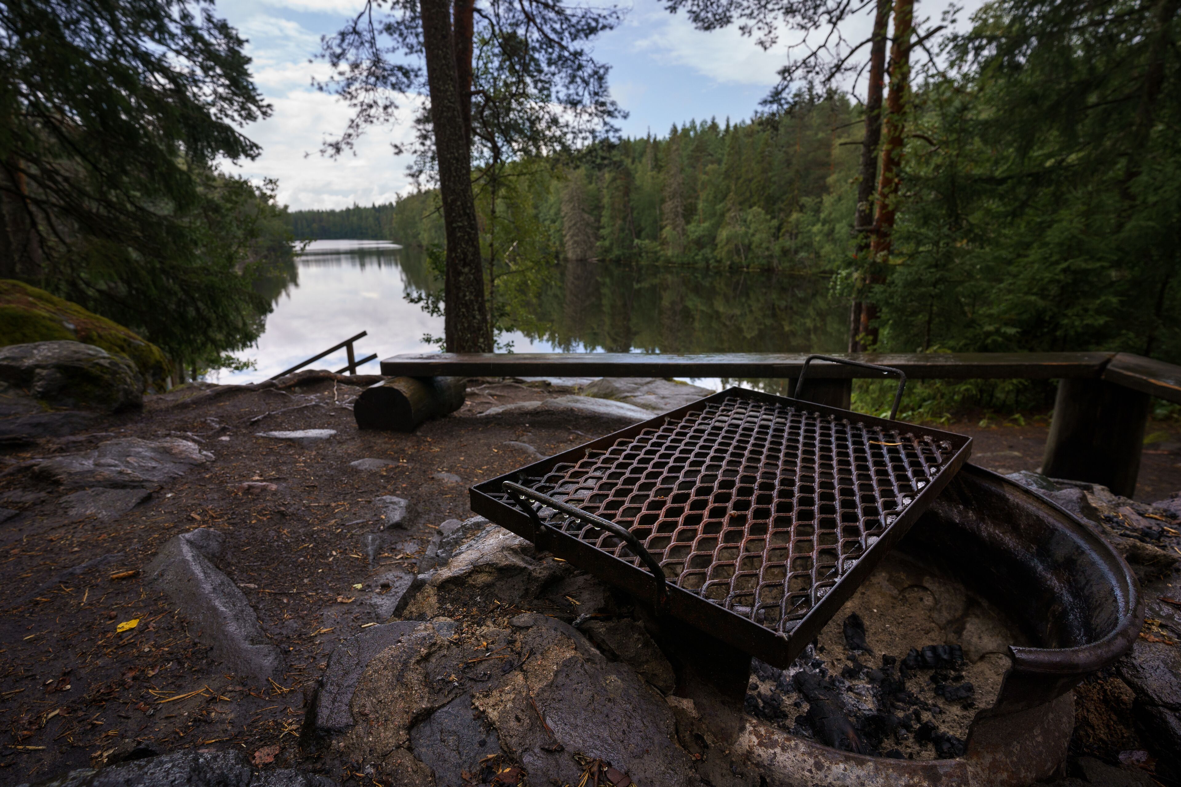 A metal grill over a campfire surrounded by rocks and trees near a lake.