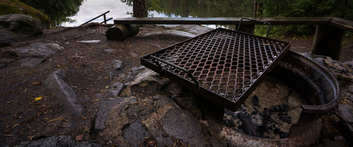 A metal grill over a campfire surrounded by rocks and trees near a lake.