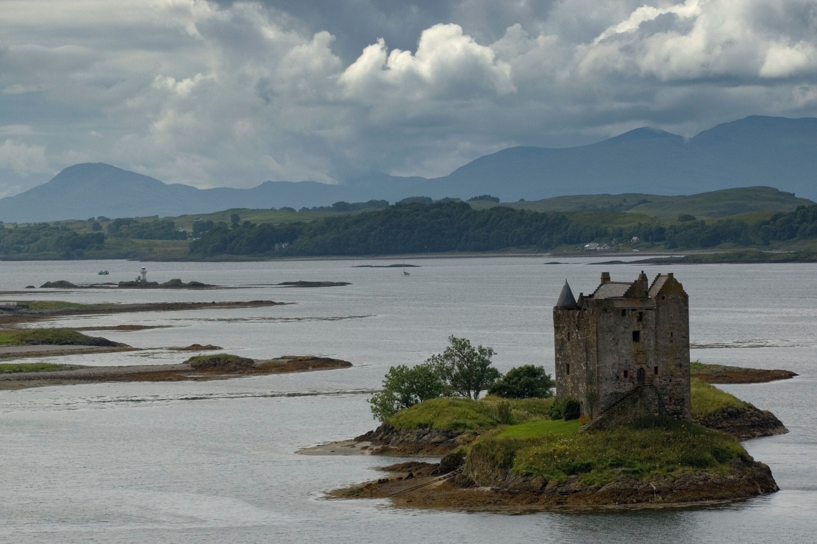 A historic stone castle on a small island surrounded by water, with rolling hills and a cloudy sky in the background.