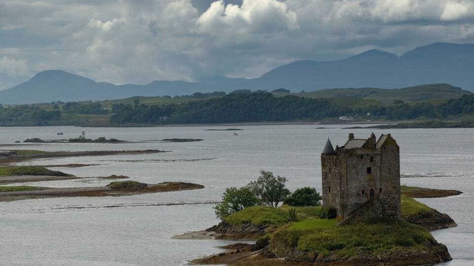 A historic stone castle on a small island surrounded by water, with rolling hills and a cloudy sky in the background.