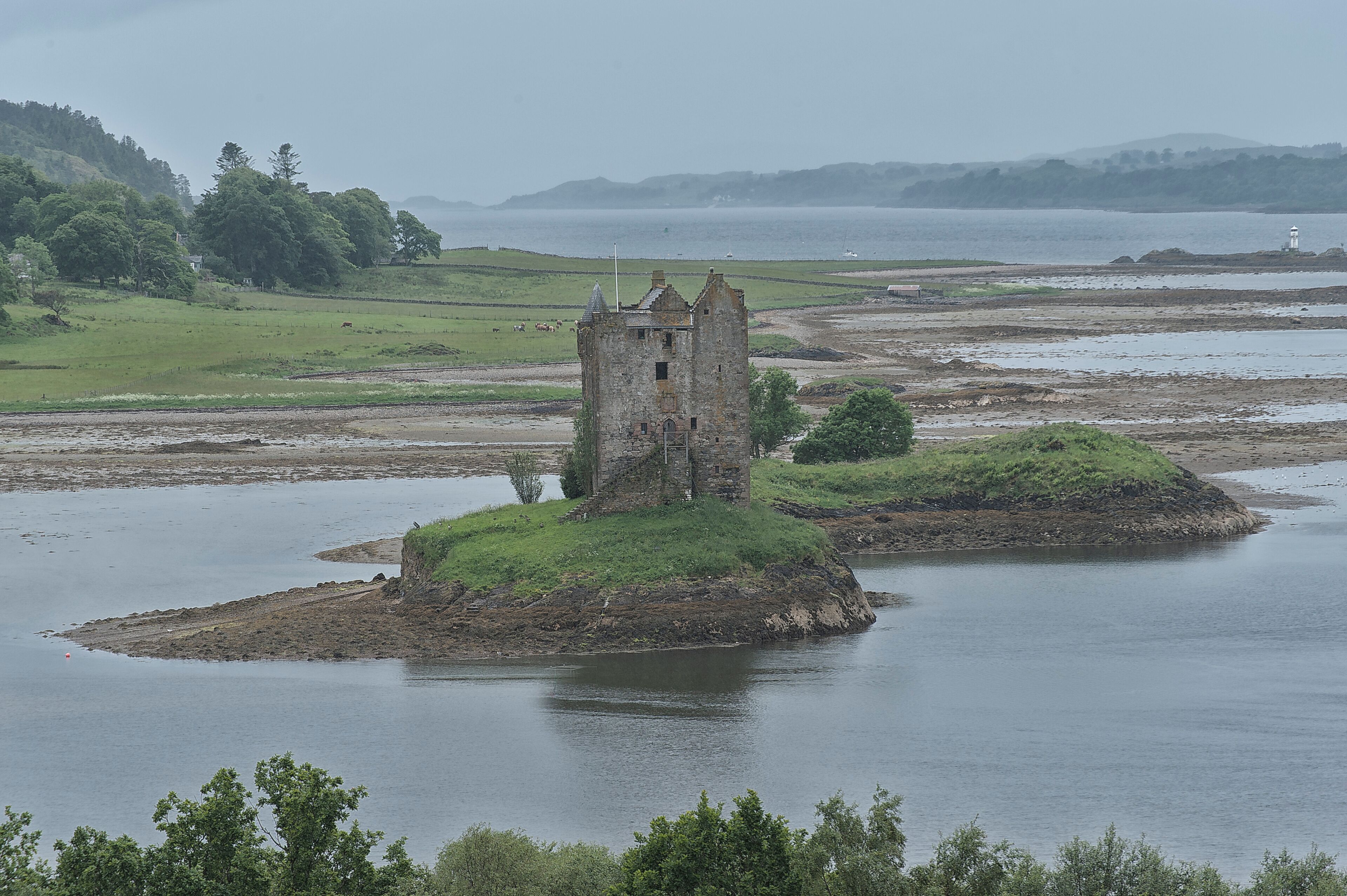 A historic tower on a small island surrounded by water, with a lighthouse in the distance.