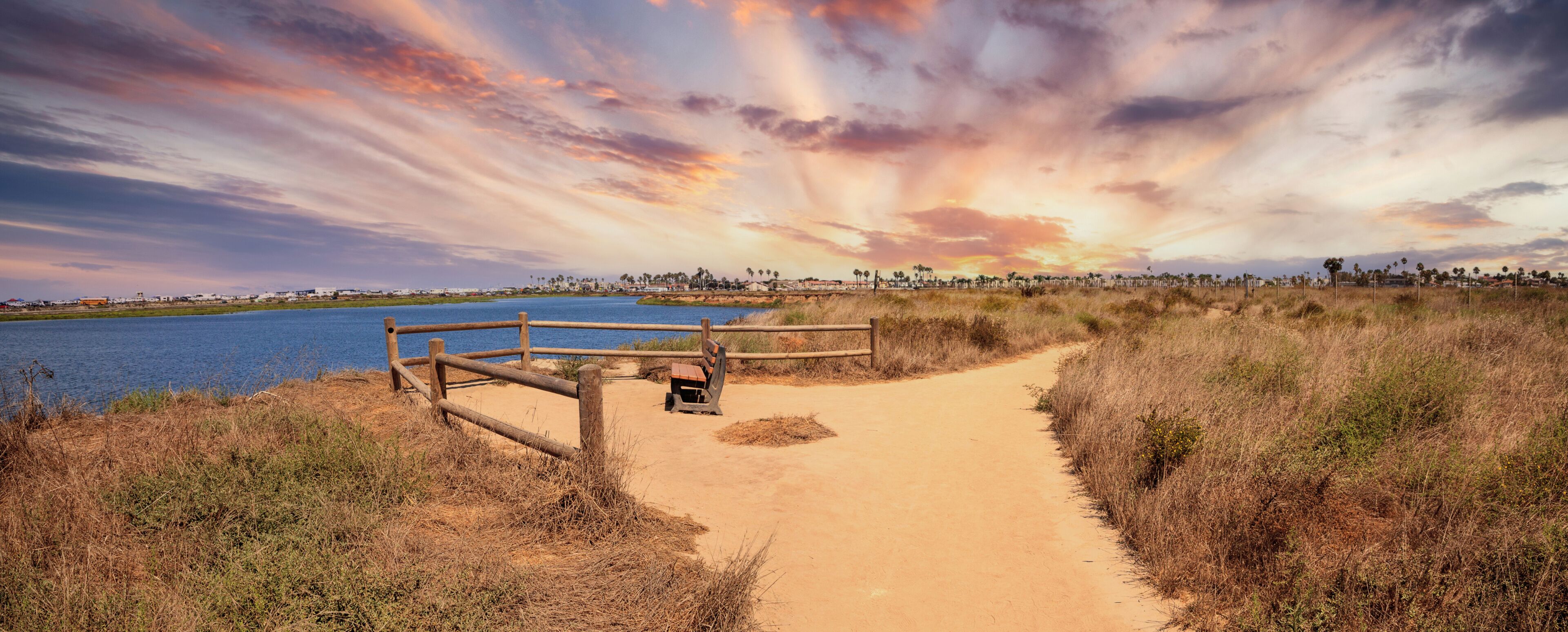 A sandy path leading to a body of water, with a wooden fence and bench, under a colorful sunset sky.