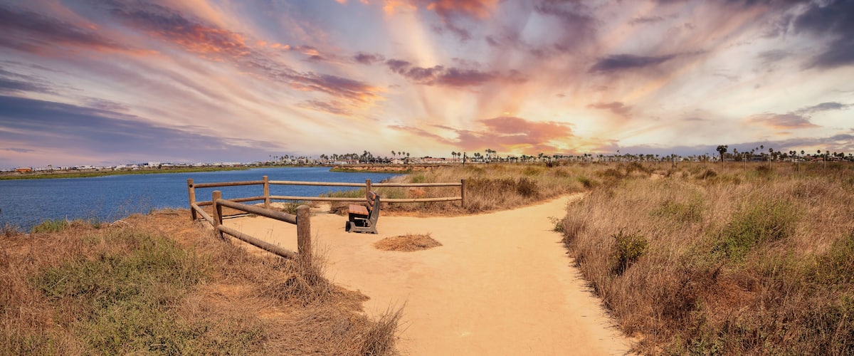 A sandy path leading to a body of water, with a wooden fence and bench, under a colorful sunset sky.