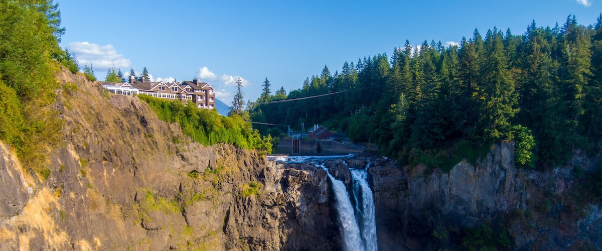 A waterfall cascading down a rocky cliff into a river, surrounded by dense forest and a building on a hill.